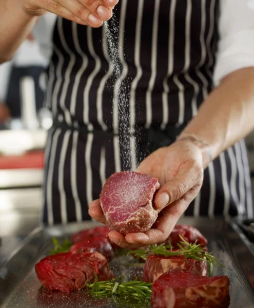 Chef with Dark Blue and White striped apron seasoning steaks on a plate with salt